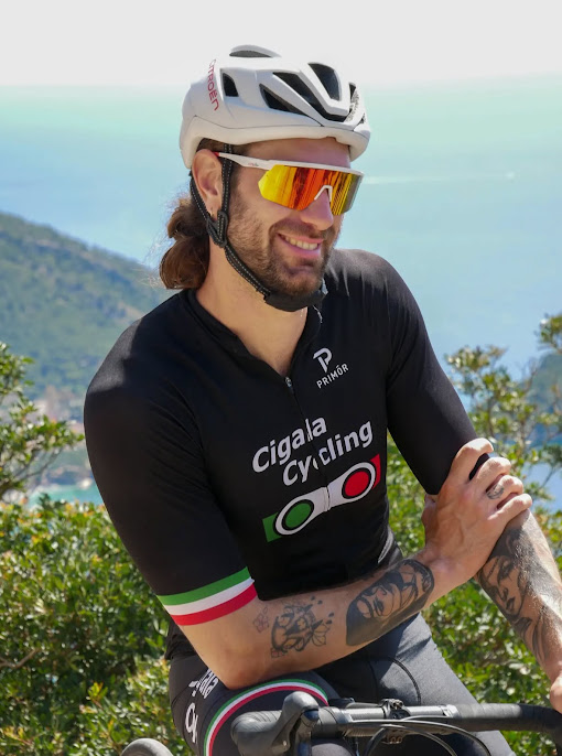 A smiling male cyclist in a black Cigala Cycling jersey, white helmet, and mirrored sunglasses posing outdoors.